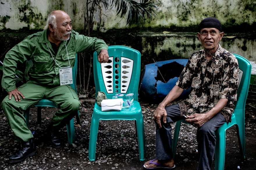 Two older men sitting on plastic chairs in conversation on a street in Indonesia, one wearing a batik shirt.