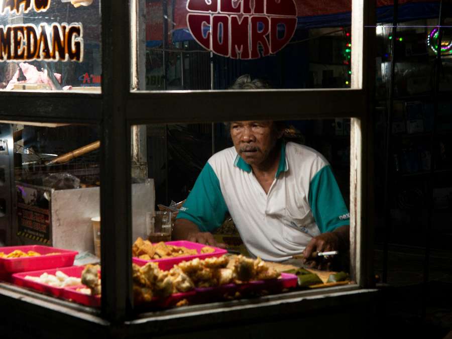 A vendor standing behind the glass case of a lit food cart on an Indonesian street at night.